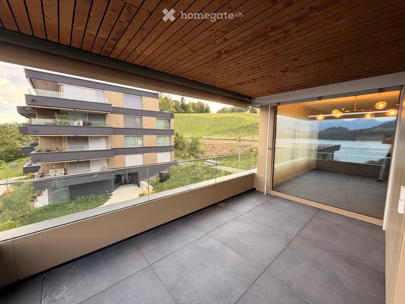 Modern balcony with wooden ceiling, glass railing, and expansive view of a lake and surrounding landscape.