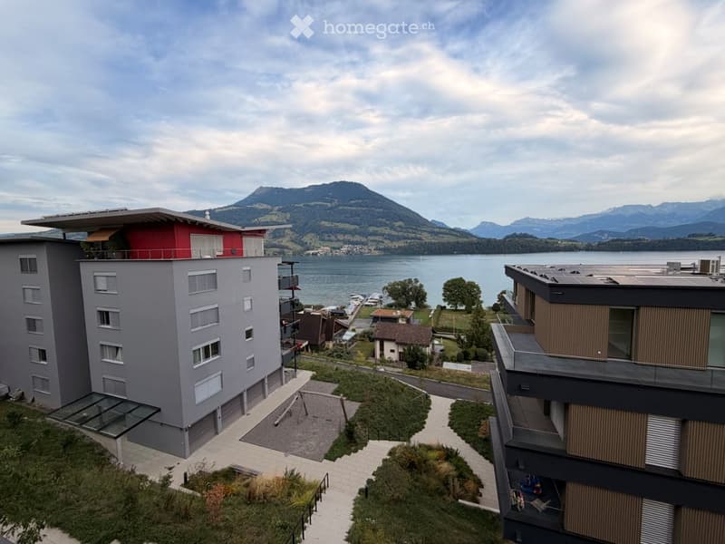 Modern multi-story residential building with a red and grey facade, overlooking a lake and mountain range. Features include balconies, a paved walkway, and surrounding greenery.