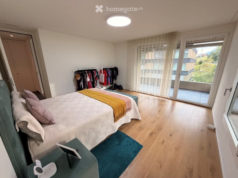 Modern bedroom with a large bed, nightstand, and a view through large windows. The room features wooden flooring, a clothing rack, and a small table with a tablet and other items.
