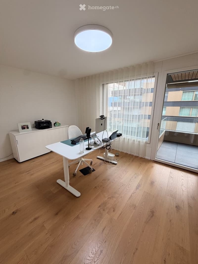 Modern office space with wooden flooring, large window with sheer curtains, round ceiling light, and a white desk with a chair, laptop, and printer.