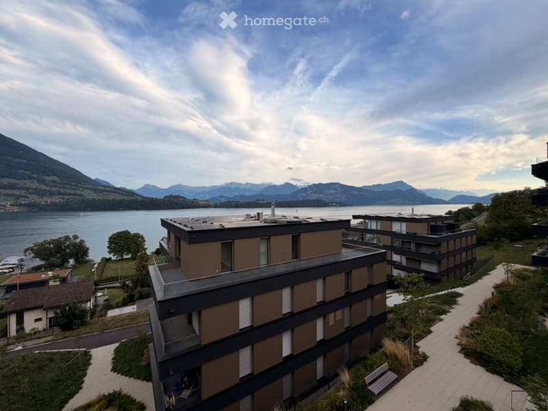 Modern multi-story building with a sleek design, featuring large windows and a balcony overlooking a scenic lake and mountain range.