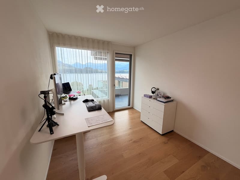 Bright, modern workspace with a large window offering a scenic mountain view. The room features a white desk with a computer setup, a tripod, and a small drawer unit holding books and accessories.