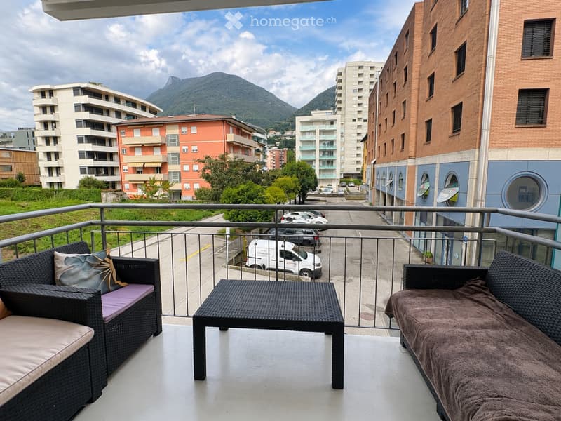 Balcony with wicker furniture, including a sofa, table, and chair, overlooking a cityscape with buildings and mountains.