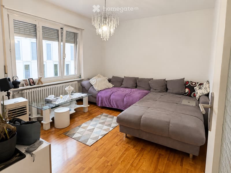 Modern living room with a grey sectional sofa, purple throw, glass coffee table, laptop, and decorative items on a wooden floor.