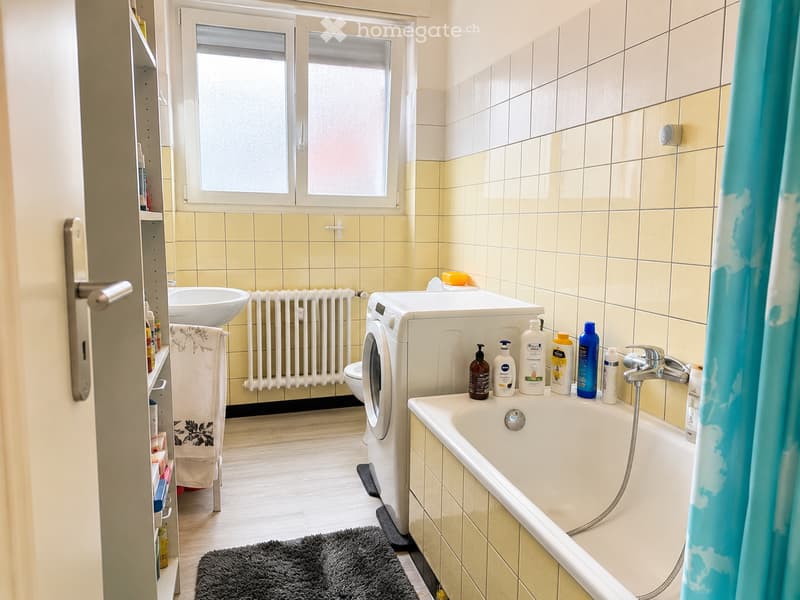 Bathroom with a bathtub, sink, washing machine, radiator, towel, and various toiletries on the edge of the tub.