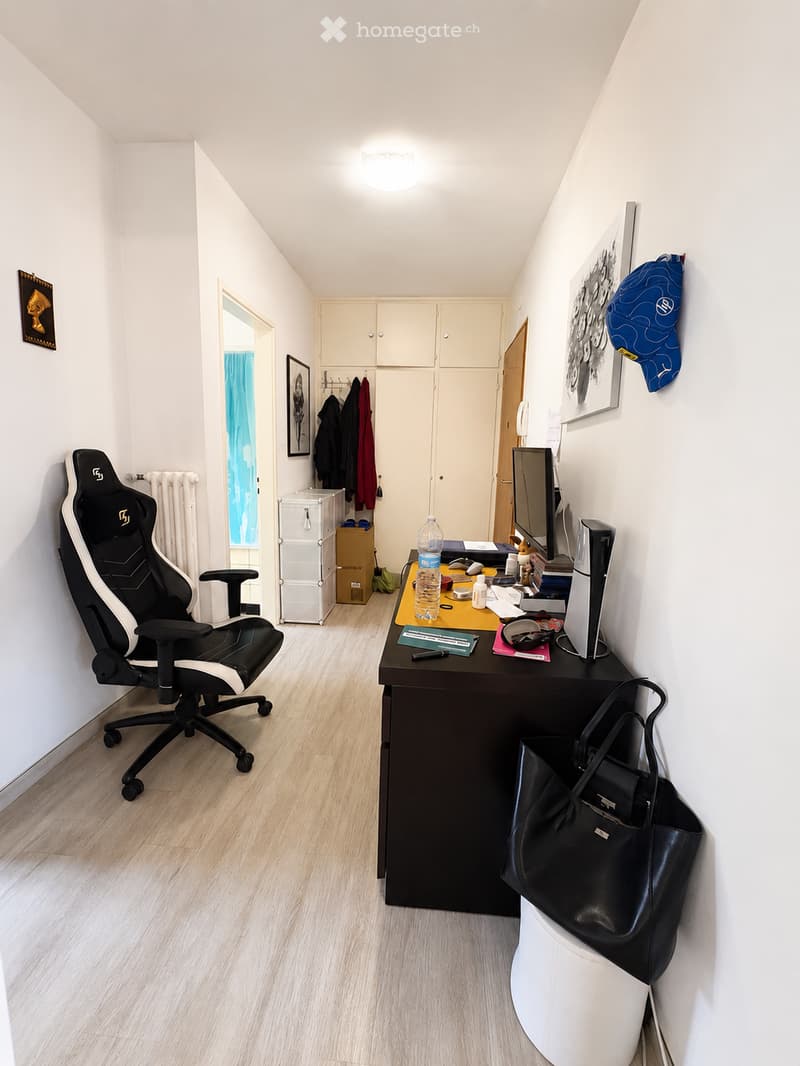 A modern, well-lit office space with a black office chair, a dark wooden desk, a computer screen, and various office supplies. The room features light wooden flooring, white walls, and built-in cabinets.