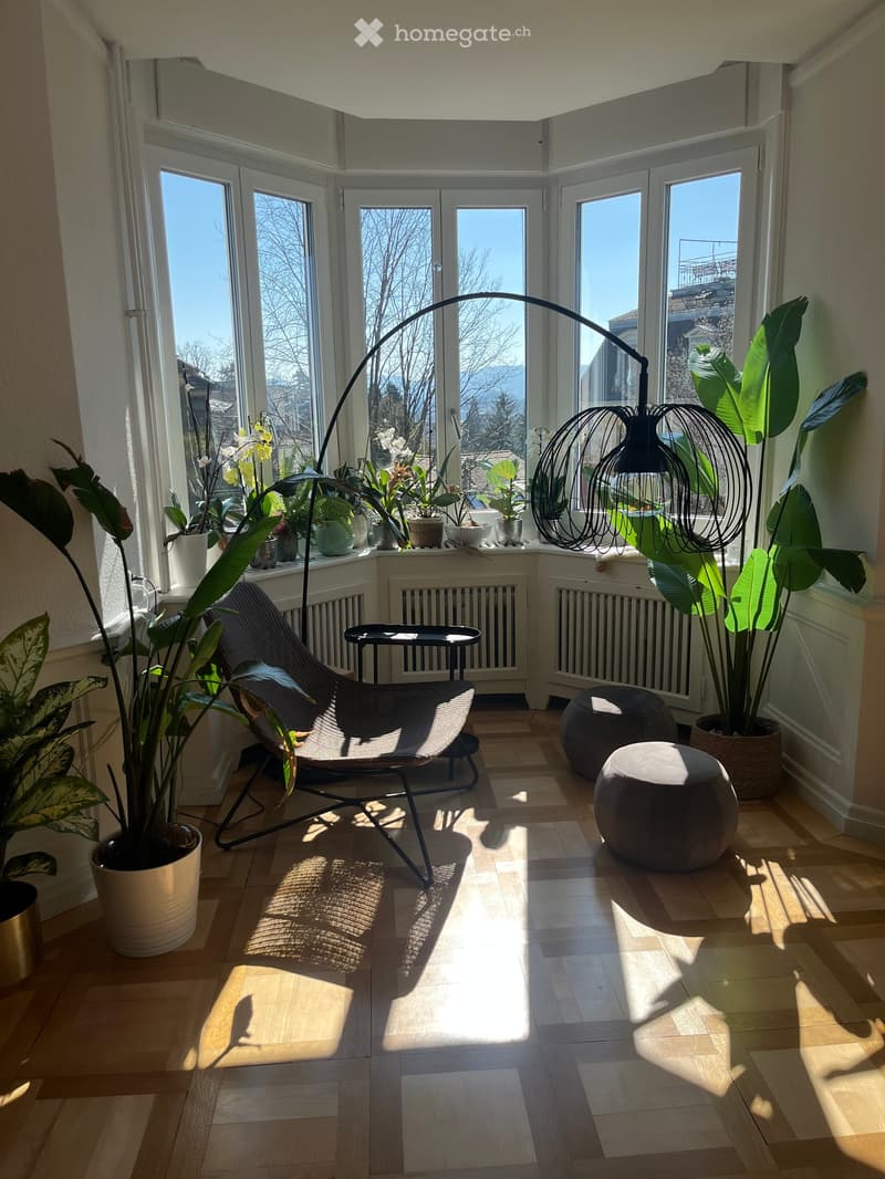 Bright sunroom with large windows, wooden floor, modern furniture including a lounge chair, two stools, a small table, and a floor lamp, surrounded by potted plants.