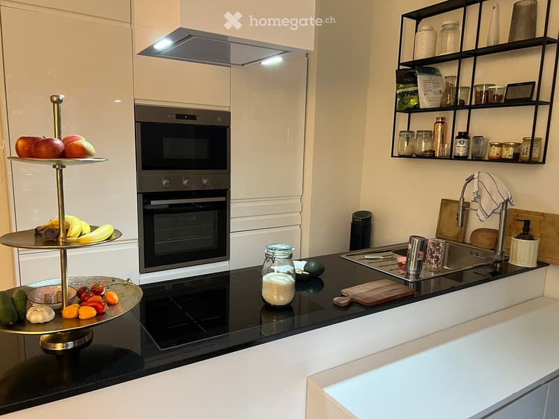 Modern kitchen with white cabinets, black countertops, stainless steel appliances, and a double-tiered fruit and vegetable display on the counter.