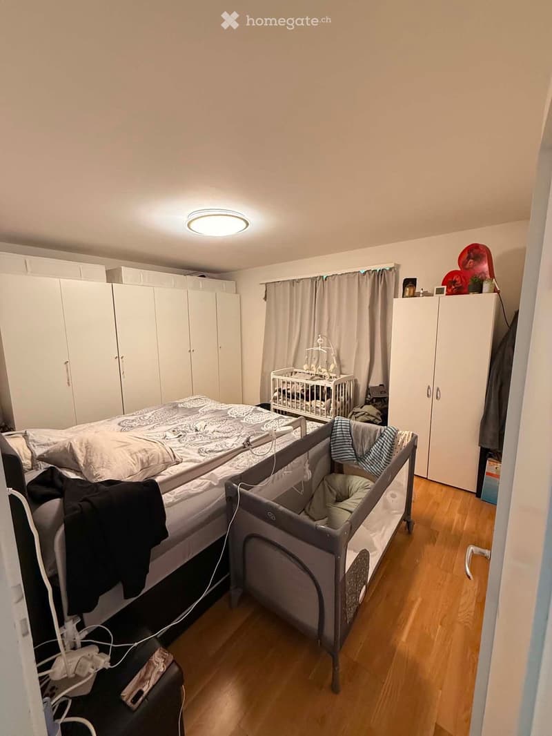 A well-lit bedroom with a double bed, a baby crib, a playpen, and built-in wardrobes. The room features wooden flooring and a ceiling light.