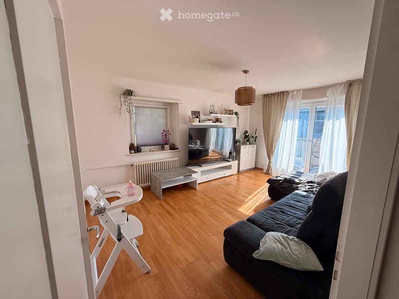 A modern living room with wooden flooring, a dark blue sofa, a coffee table, a baby chair, a television, and a large window with curtains leading to a balcony.