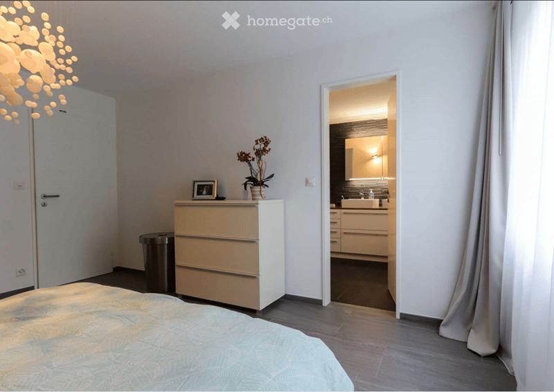 white walls, grey floor, double bed, dresser with potted plant, trash can, lamp, door to another room
