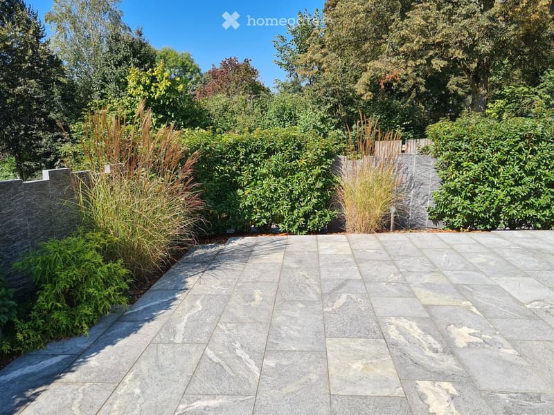 A paved courtyard area surrounded by stone walls, greenery, and trees. The paving stones are large and gray with varying textures.