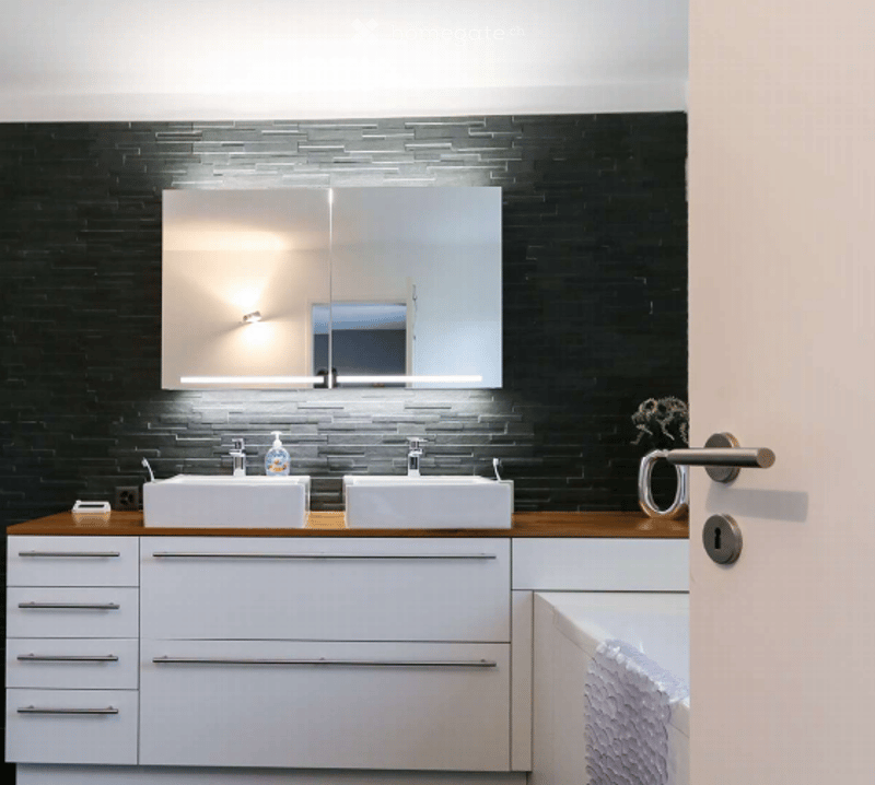 White cabinets, black tiled wall, mirror with built-in light, wooden countertop, white bathtub, two sinks, faucets, shower area, potted plant on countertop