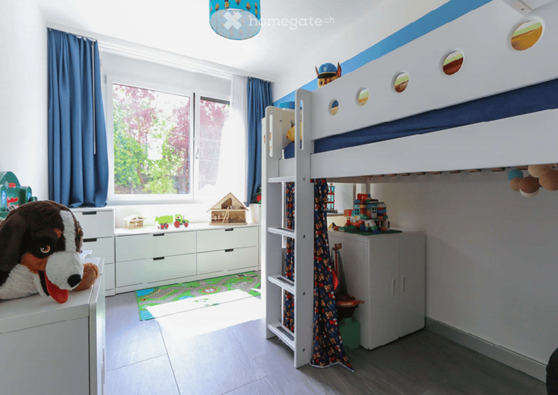Modern children's room with a loft bed, white walls, blue curtains, white wardrobe, playmat, window, soft toys, and toys on shelves.