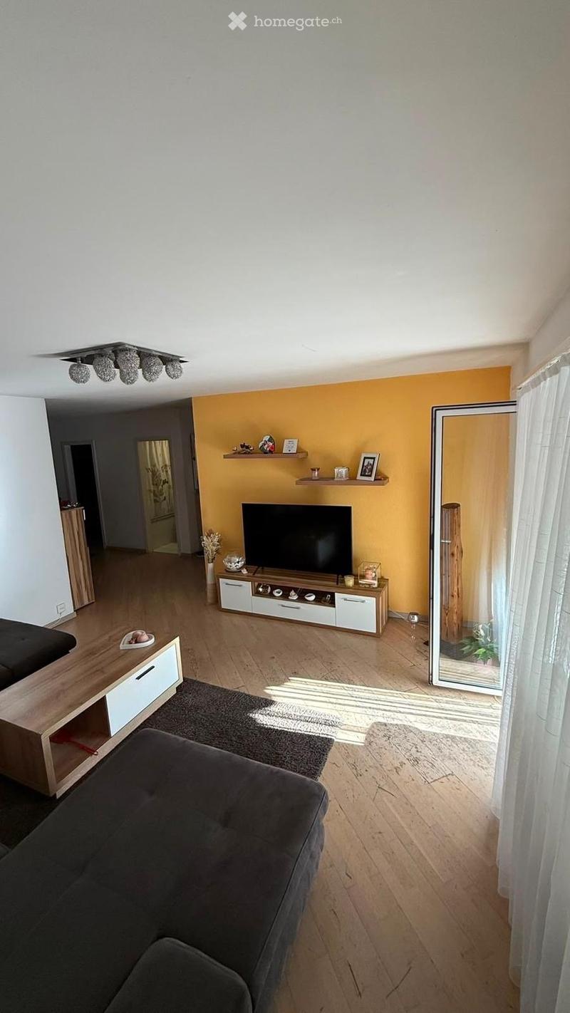 Modern living room with wooden flooring, grey sofa, coffee table, TV stand, shelves, and large mirror with curtain.