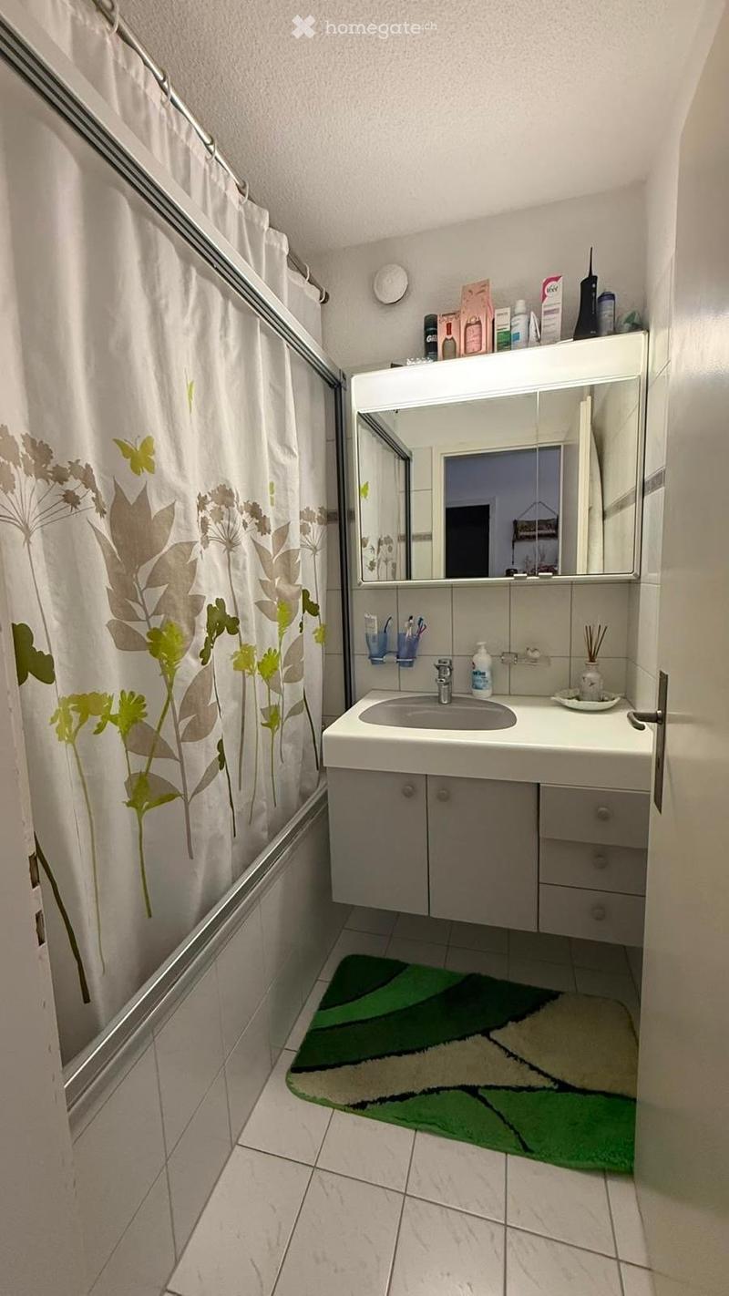 A compact bathroom with a white sink, mirror, cabinet, and a shower area with a floral-patterned curtain. A green and white rug is placed on the tiled floor.