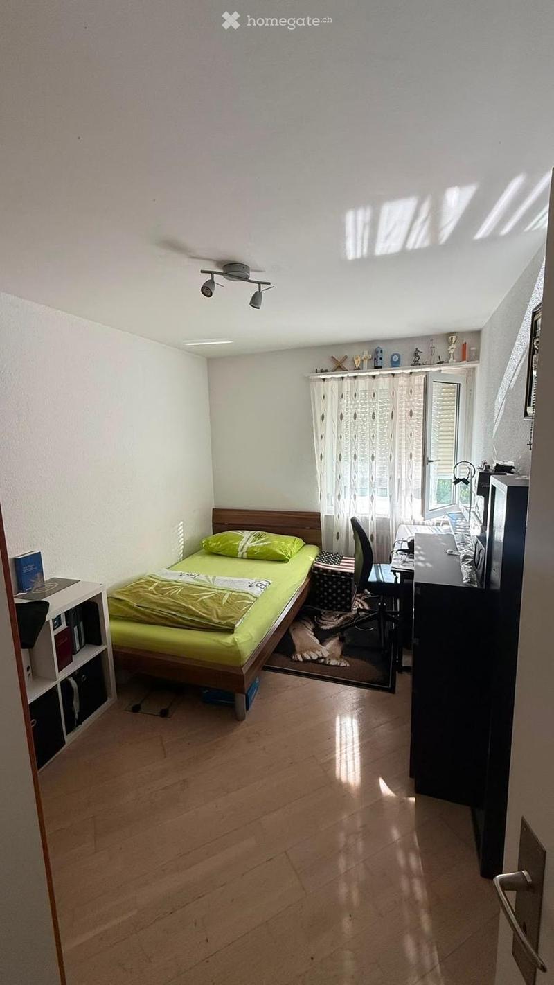 A small, well-lit bedroom with a single bed, a desk, a bookshelf, and a cat. The room features wooden flooring and white walls.