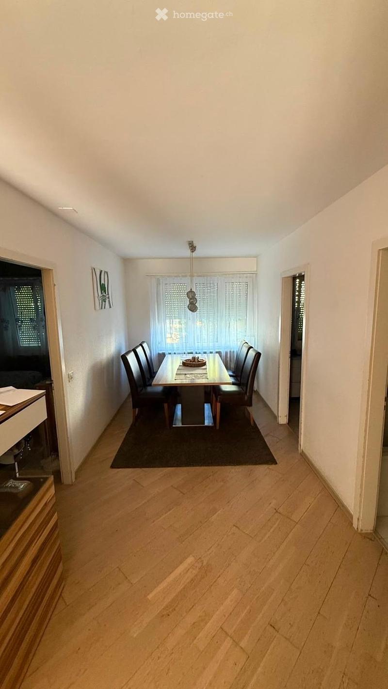 Dining room with wooden floor, dining table, chairs, and a hanging light fixture. There is a window with curtains and a picture frame on the wall.
