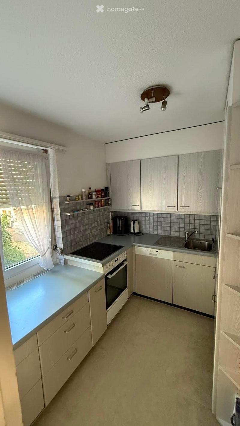 Modern kitchen with light-colored cabinets, stainless steel appliances, and a window with blinds. Features an oven, sink, dishwasher, and countertop with a coffee maker and small items on a shelf.