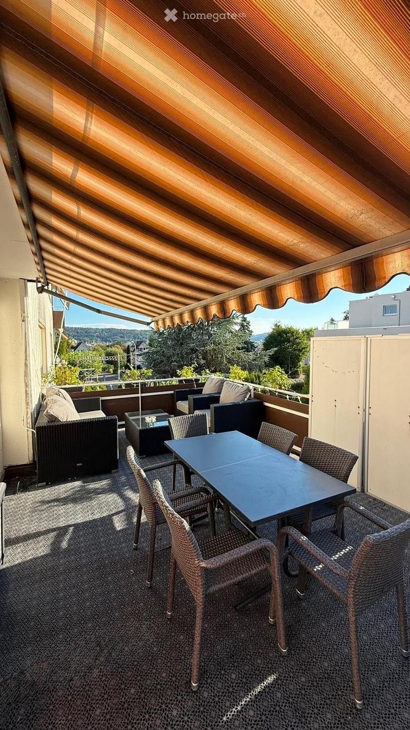 Balcony with a striped awning, a dining table with chairs, and a sofa set, overlooking a scenic view.