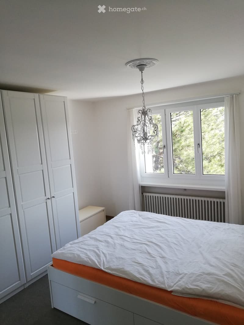 Bright bedroom with a white bed, white wardrobe, chandelier, and large windows with white curtains.
