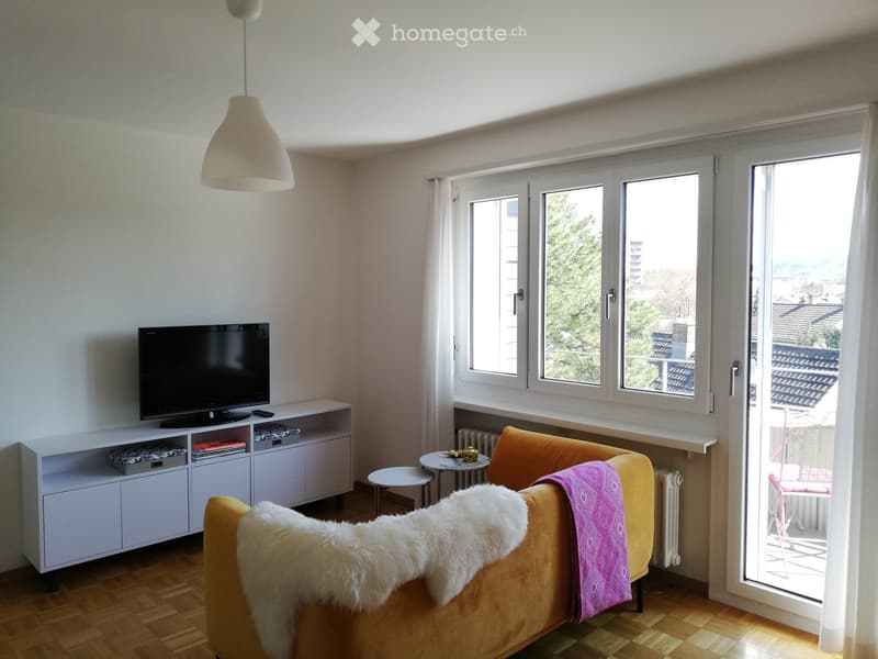 Living room with a yellow sofa, a small round table, a television on a white cabinet, and a hanging lamp. The room has large windows with a view of the city and a balcony accessible through a sliding glass door.