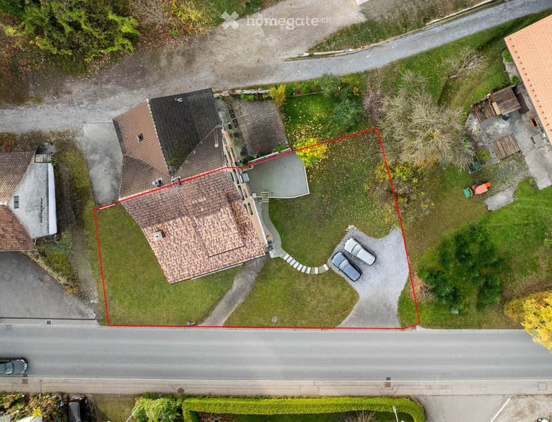 Aerial view of a house with a garage, driveway, and two parked cars, surrounded by a grassy area and trees. Red lines outline the property boundaries.