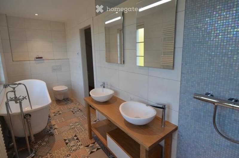 Modern bathroom with a white bathtub, toilet, two sinks on a wooden vanity, and large mirrors above the sinks. The walls are tiled in white and blue, and the floor has a patterned tile design.