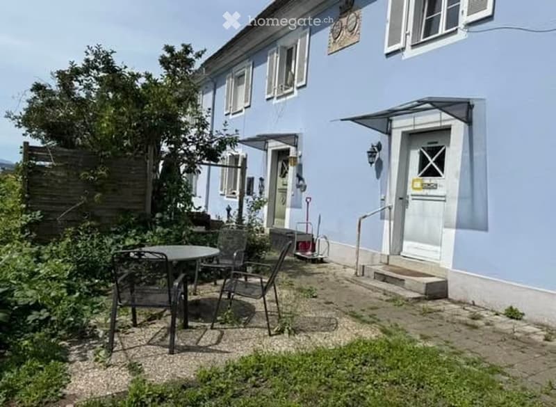 A small patio area with a round table and four chairs, adjacent to a light blue house with white-framed windows and a small entrance.