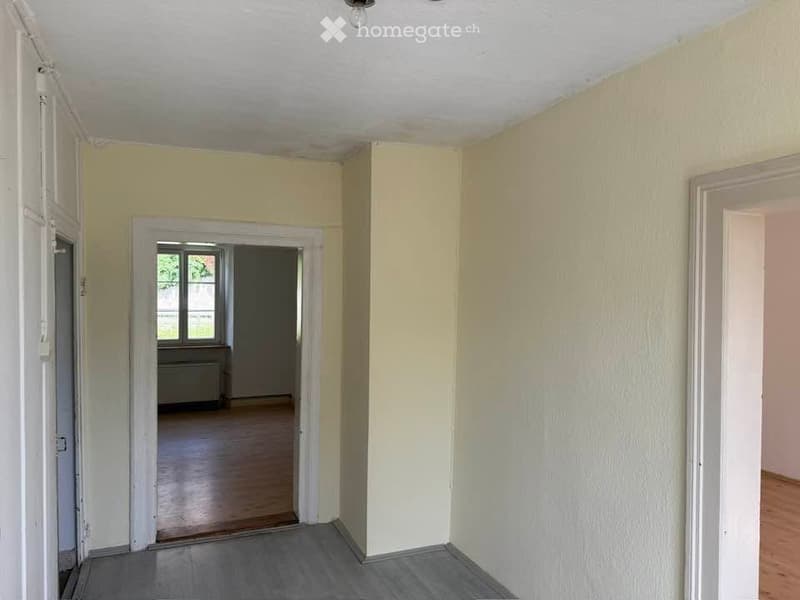 Empty hallway with light yellow walls, wooden flooring, and a doorway leading to another room with a window and radiator.