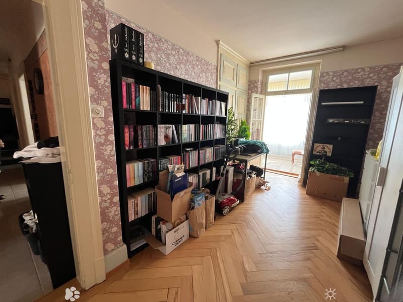 A room with wooden flooring, a large bookshelf filled with books, a small table with a plant, and a vacuum cleaner. There is also a refrigerator and some boxes in the room.