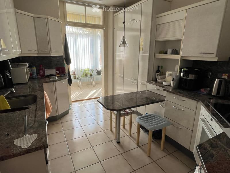 Modern kitchen with white cabinets, granite countertops, and a central island. Appliances include a coffee maker, microwave, and oven. Stools are placed around the island, and a small table with chairs is visible in the adjacent area.