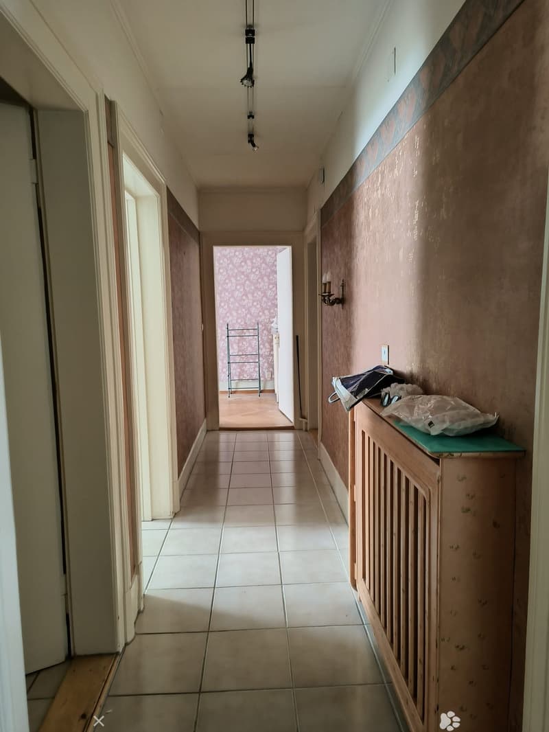 Empty hallway with tiled floor, wooden walls, and a radiator on the right side. A small green surface holds a few items, and a doorway leads to another room with floral wallpaper.