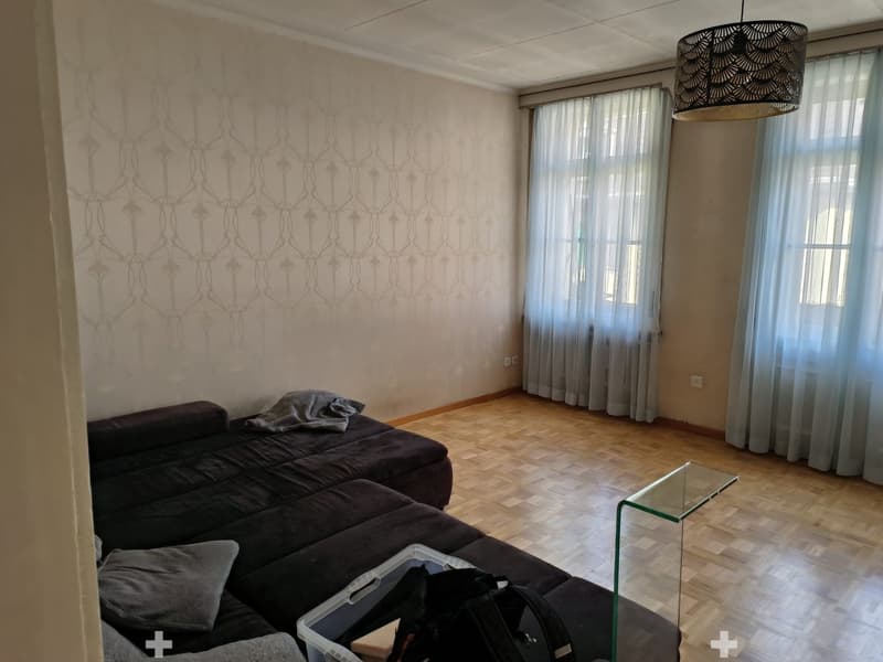 Modern living room with dark grey sectional sofa, glass coffee table, and large windows with sheer curtains. Wooden floor and patterned wallpaper.