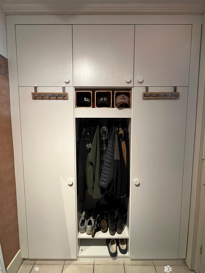 A modern hallway closet with white cabinetry, multiple hanging rods for coats and jackets, and a lower shelf for shoes and boots. The closet features built-in storage compartments and a coat rack on the left side.