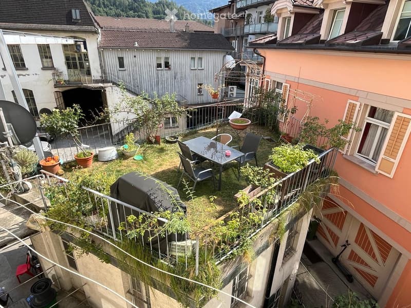 A small, fenced garden area with a table, chairs, potted plants, and a covered grill. The garden is surrounded by neighboring buildings and has a metal railing.