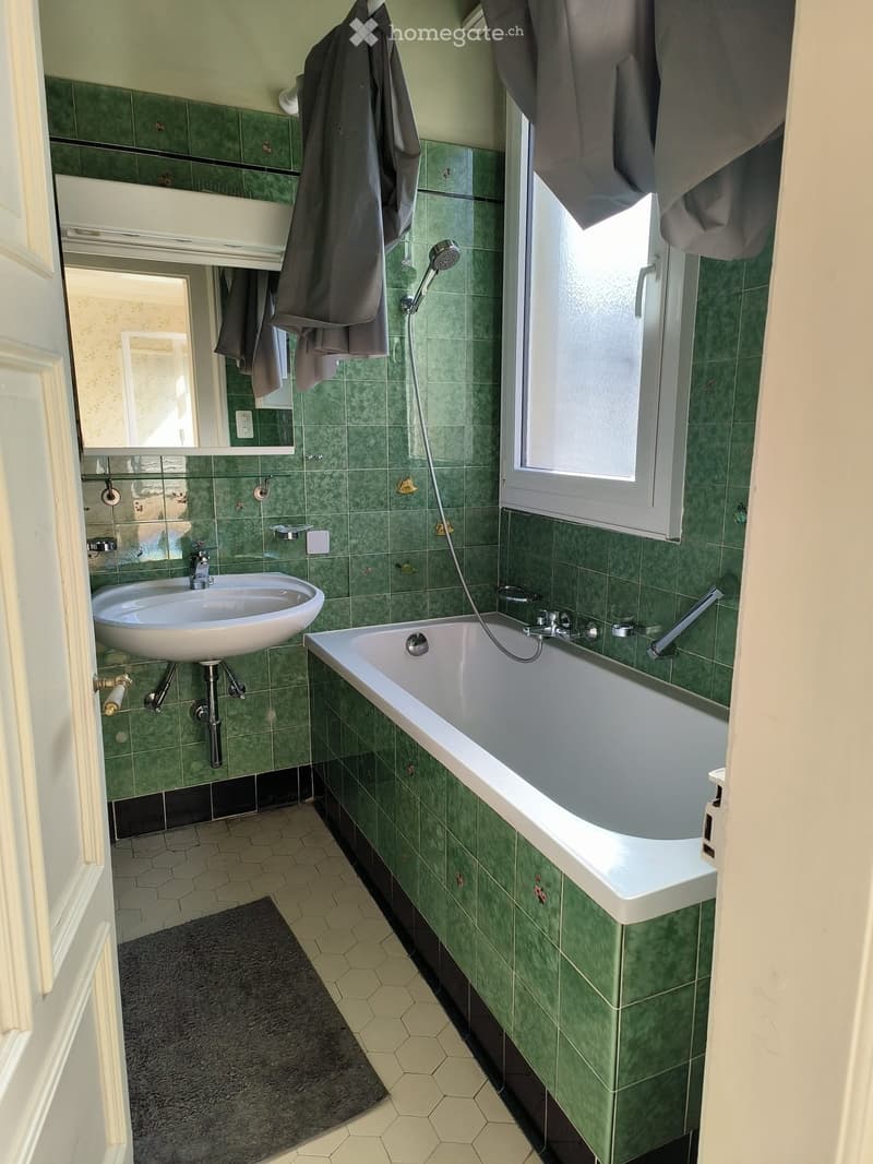 Bathroom with green tiled walls, a bathtub, a sink, a mirror, and a window with curtains.