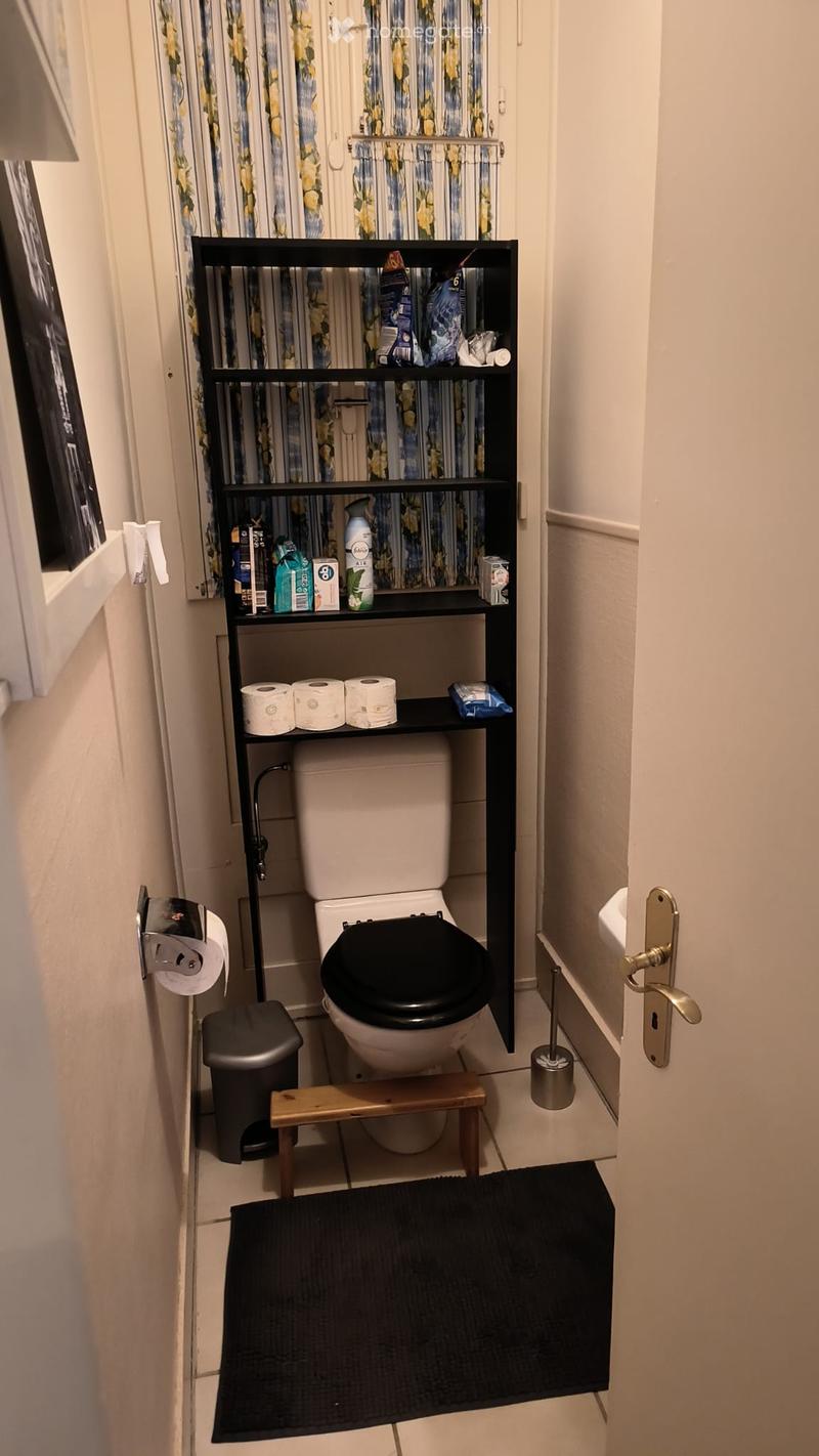 Compact bathroom with a toilet, black shelf holding toiletries, toilet paper, a small wooden stool, a black rug, and a trash can.