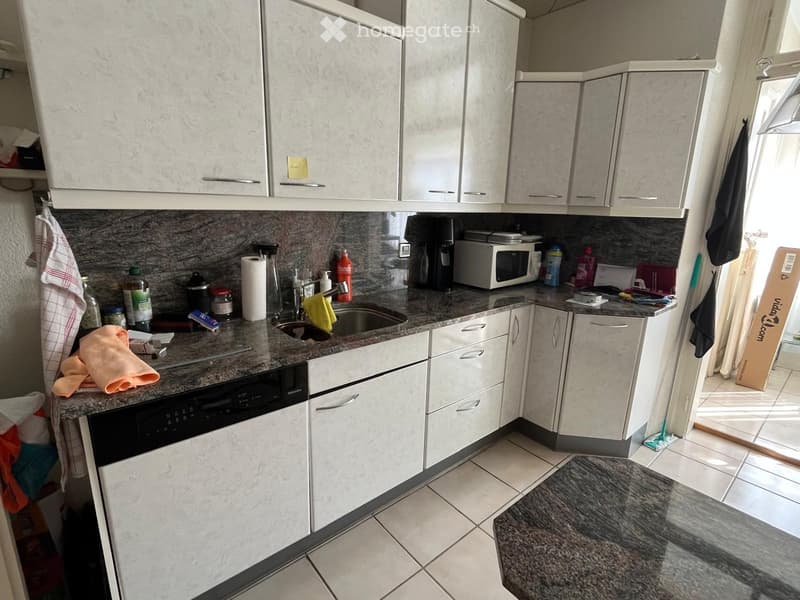Modern kitchen with white cabinets, granite countertops, and stainless steel appliances including a dishwasher and microwave. A sink with a faucet is visible, along with various items on the counter such as a coffee maker, bottles, and a towel.