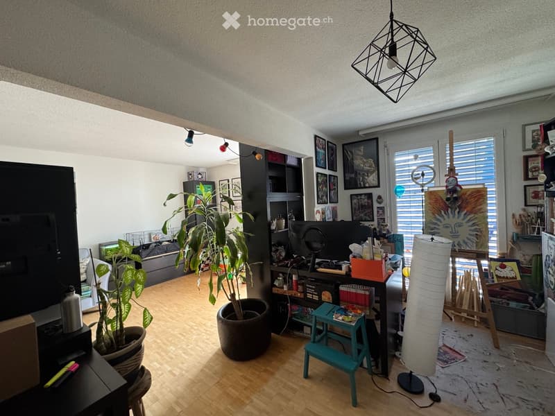 The room features a wooden floor, large windows with blinds, a hanging geometric lamp, a sofa, a TV screen, a shelf with various items, a tall plant in a pot, a small teal stool, and a tall white lamp.
