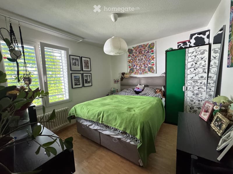 Cozy bedroom with a green duvet, wooden floor, large windows, nightstand, and decorative artwork.