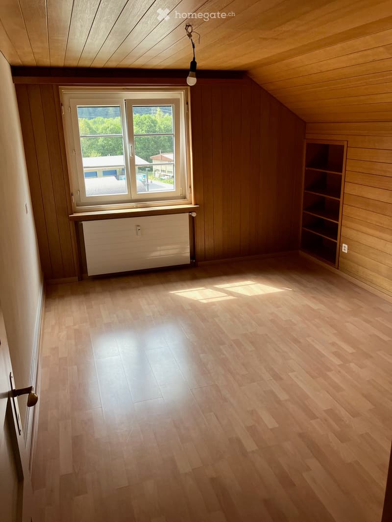 Empty attic room with wooden walls, wooden floor, large window, radiator, and built-in shelving unit.