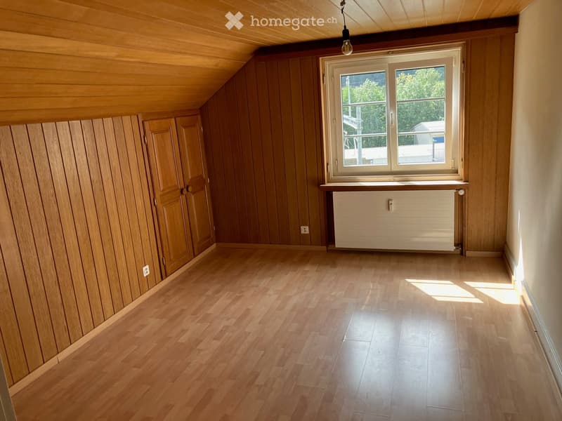 Empty attic room with wooden walls, wooden floor, large window, radiator, and exposed wooden ceiling beams.