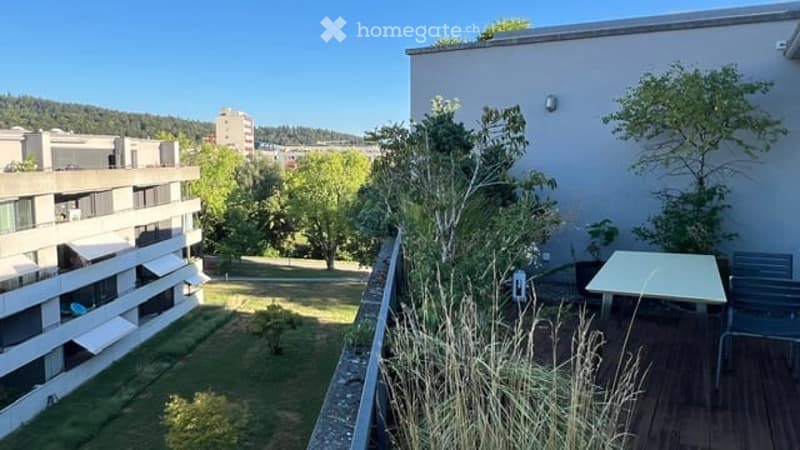 Balcony with wooden decking, small table and chairs, overlooking a green park area with trees and neighboring buildings.