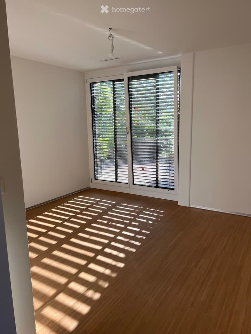 Empty room with wooden flooring and large sliding glass doors with blinds, allowing natural light to create striped shadows on the floor.