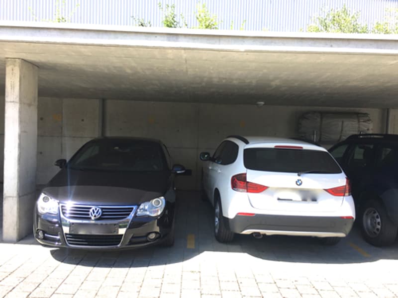 Underground parking area with two parked cars, one black and one white, under a concrete ceiling with visible support pillars.