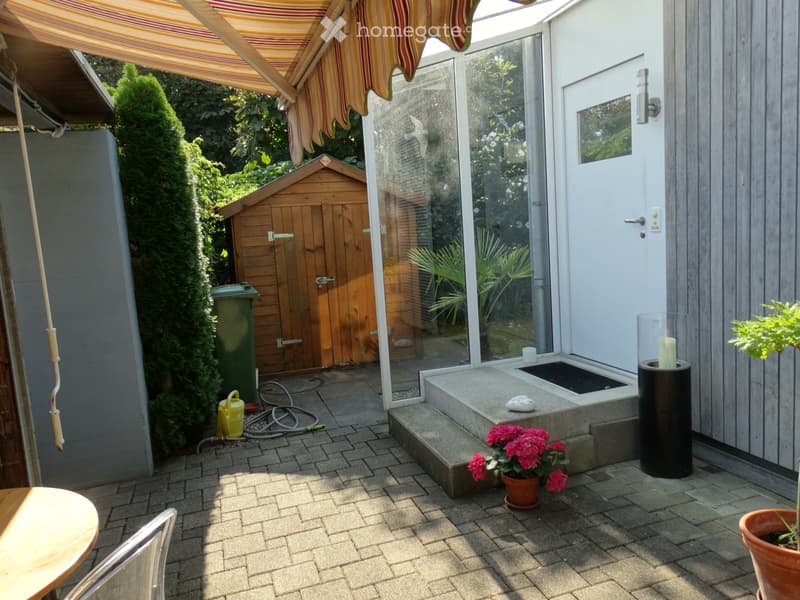 Brick patio with a table, chairs, flower pots, and a candle holder, adjacent to a wooden shed and a covered entrance.