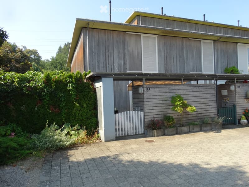 Modern two-story house with wooden exterior, large windows, and a covered entrance area. The house features a small garden with potted plants and a white picket fence.