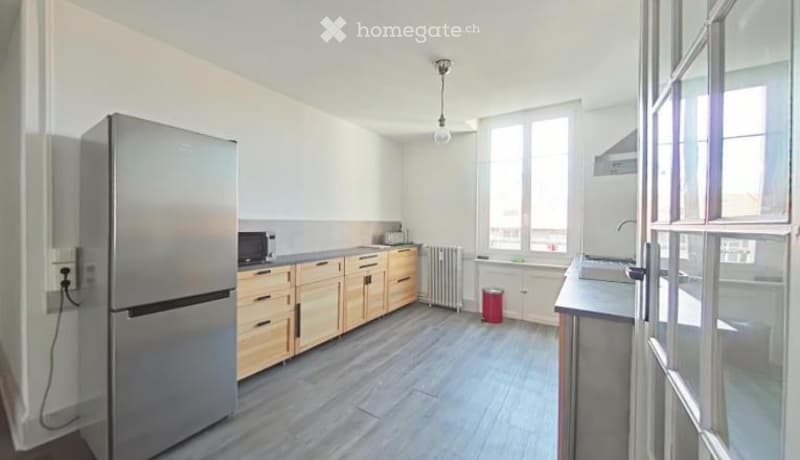 Modern kitchen with light wooden cabinets, stainless steel refrigerator, microwave, sink, and radiator. The space is well-lit with large windows and a grey tiled floor.