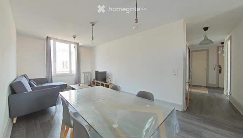 Modern, well-lit living and dining area with a grey sofa, coffee table, TV stand, and a glass dining table with chairs.